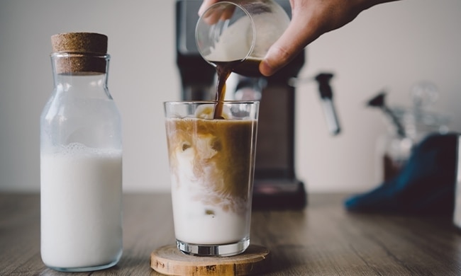 Persona che versa caffè in un bicchiere di latte messo sul tavolo con bottiglia di latte accanto