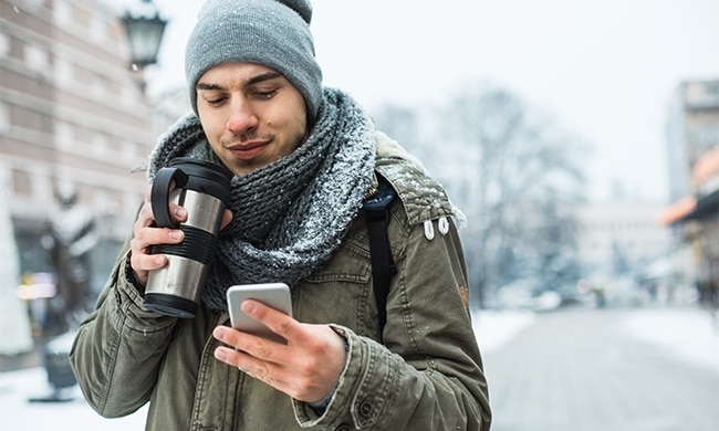 Ragazzo in città sotto la neve che beve caffè dal thermos mentre guarda il cellulare